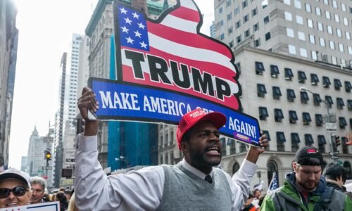 African American rally-goer wearing a Trump slogan hat boldly holds a patriotic-themed thumbs-up sign reading "Trump | Make America Great Again!"