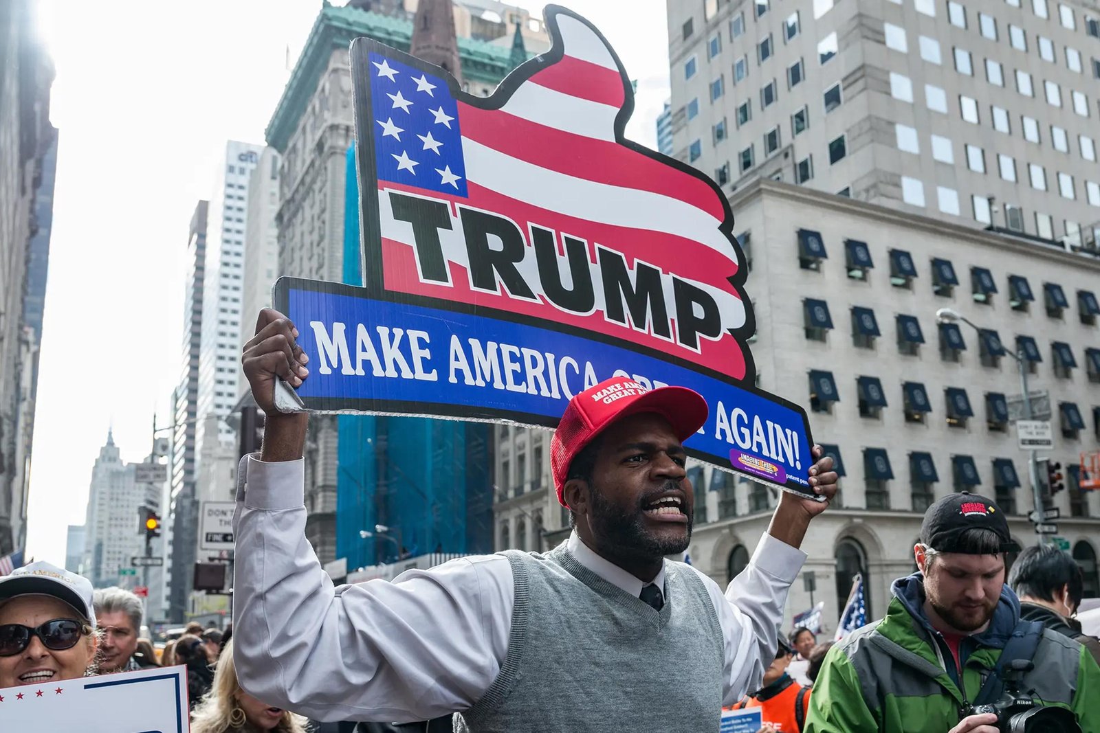 African American rally-goer wearing a Trump slogan hat boldly holds a patriotic-themed thumbs-up sign reading "Trump | Make America Great Again!"